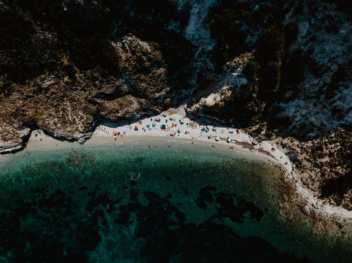 Drohnenaufnahme einer felsigen Badebucht mit bunten Sonnenschirmen am Strand und türkisfarbenem Wasser.
