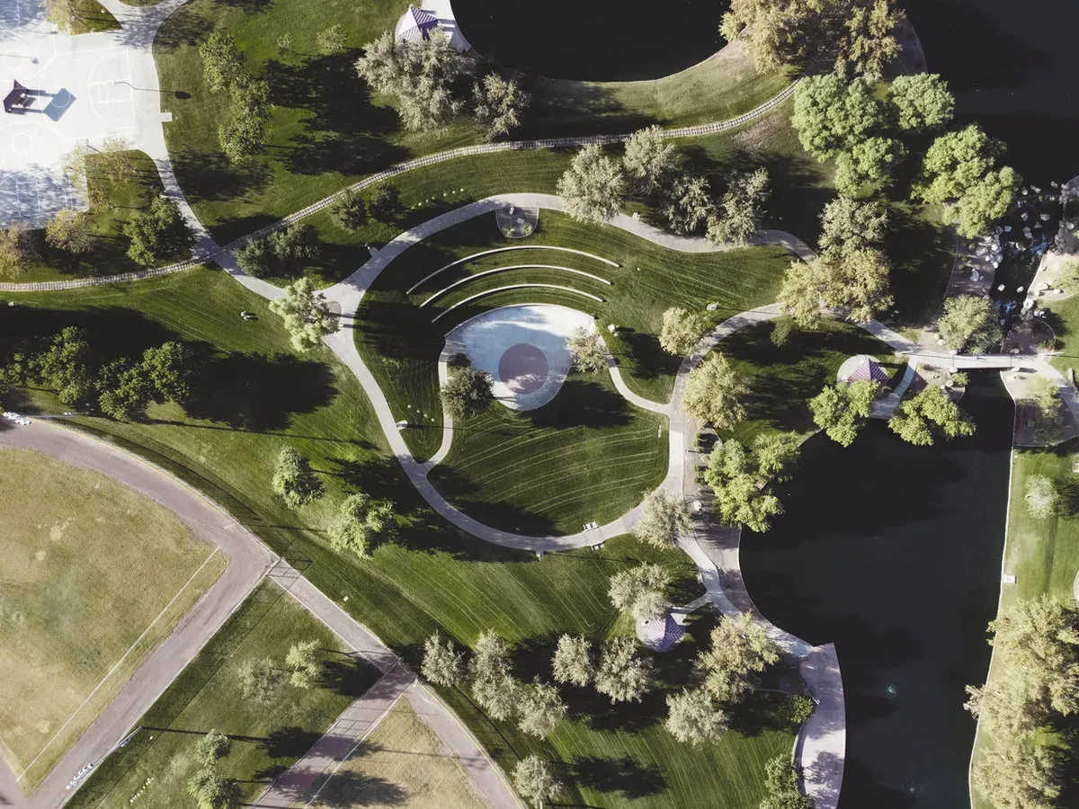 Grüne Parkanlage aus der Luft mit geschwungenen Wegen, einem runden Wasserbecken und dunklen Teichen.