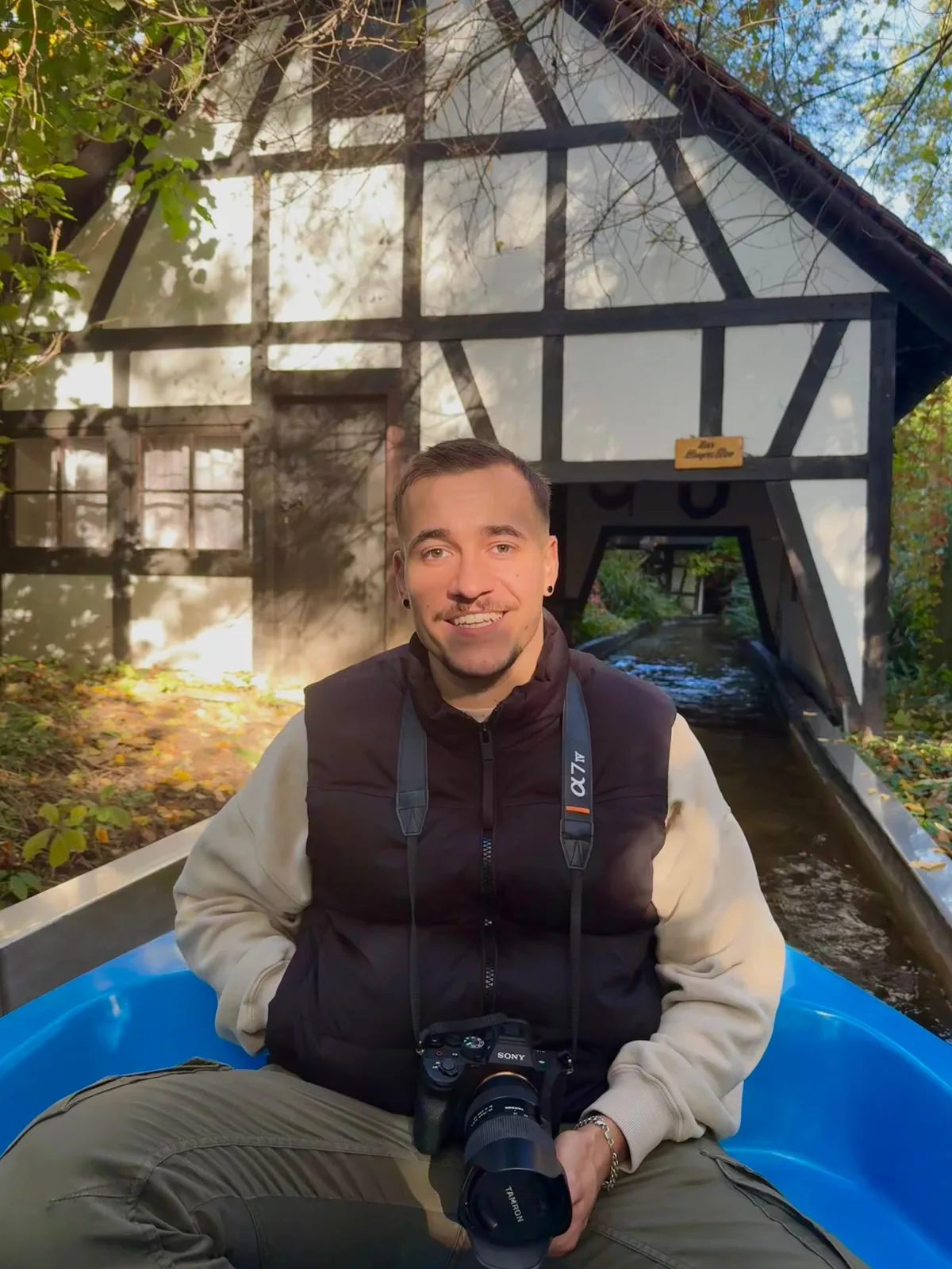 Luca Keicher sitzt mit Kamera in einem blauen Boot, dahinter ein Fachwerkhaus mit Wasserlauf.