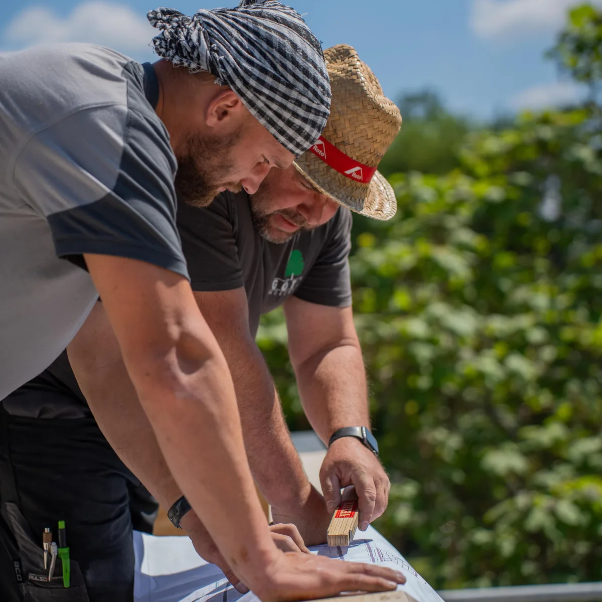Zwei Handwerker beugen sich im Sonnenlicht über einen Bauplan, einer misst mit dem Zollstock nach.