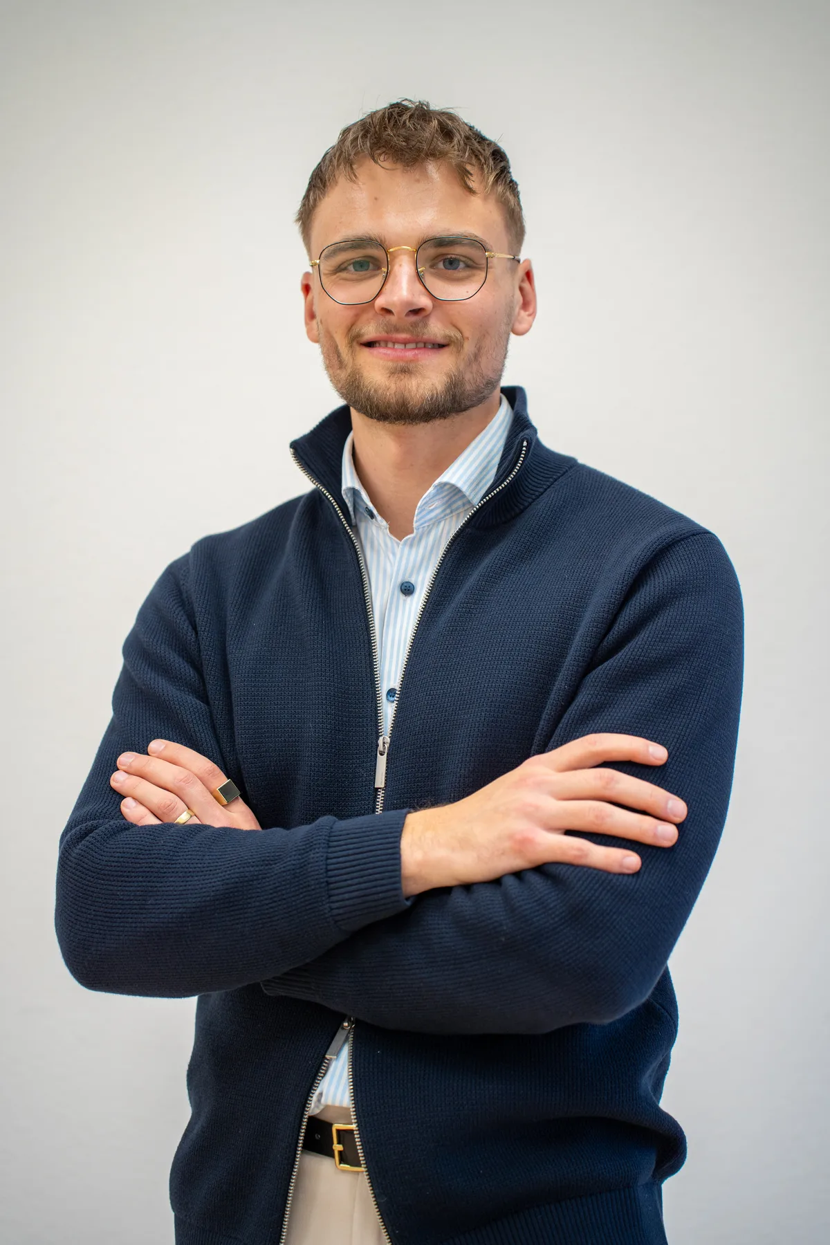 Businessportrait eines jungen Mannes mit Brille und dunkelblauer Strickjacke, die Arme verschränkt.