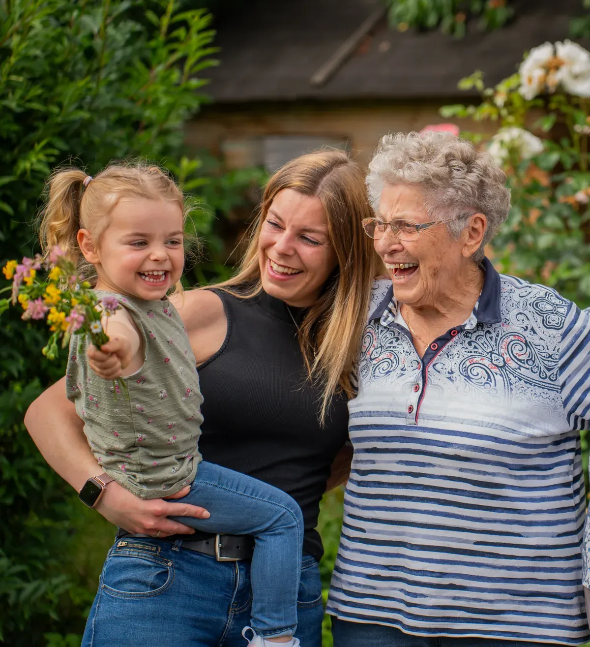 Drei Generationen lachen zusammen im Garten, das Kind hält einen bunten Blumenstrauß in der Hand.