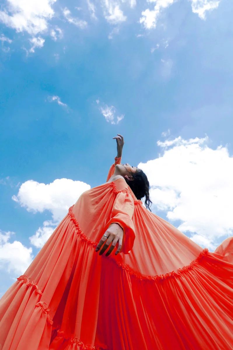 Frau in weitem orangefarbenem Kleid streckt den Arm zum Wolkenhimmel, aufgenommen aus der Froschperspektive.