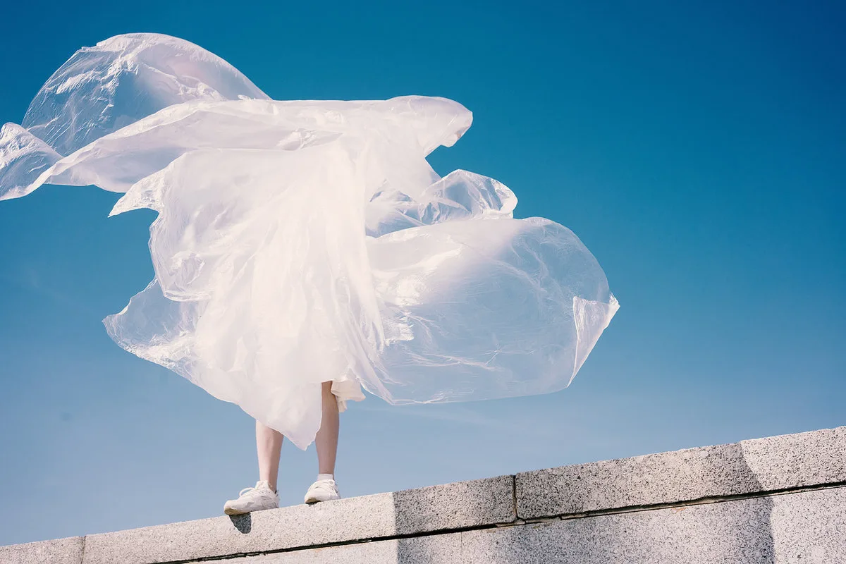 Eine Person steht auf einer Mauer, verhüllt von wehender transparenter Folie vor tiefblauem Himmel.