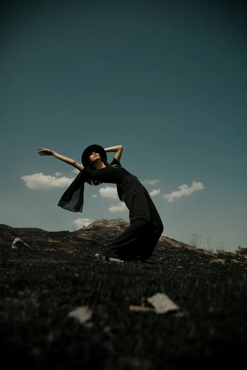 Frau in schwarzem Kleid und Hut lehnt sich in dramatischer Pose über eine dunkle Wiese vor blaugrünem Himmel.