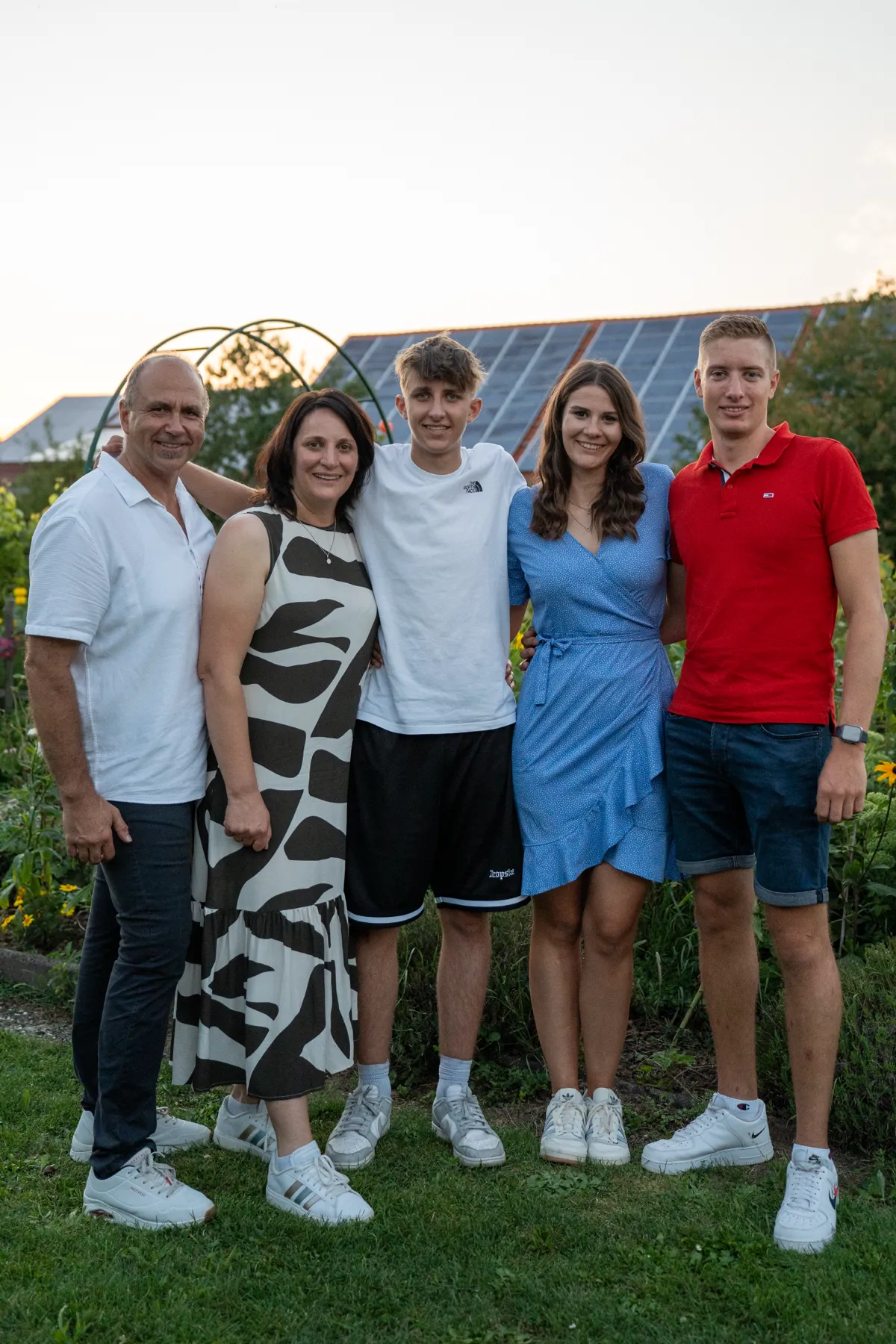 Fünf Personen posieren lächelnd im Garten, darunter eine Frau im blauen Kleid und Männer in Freizeitkleidung.