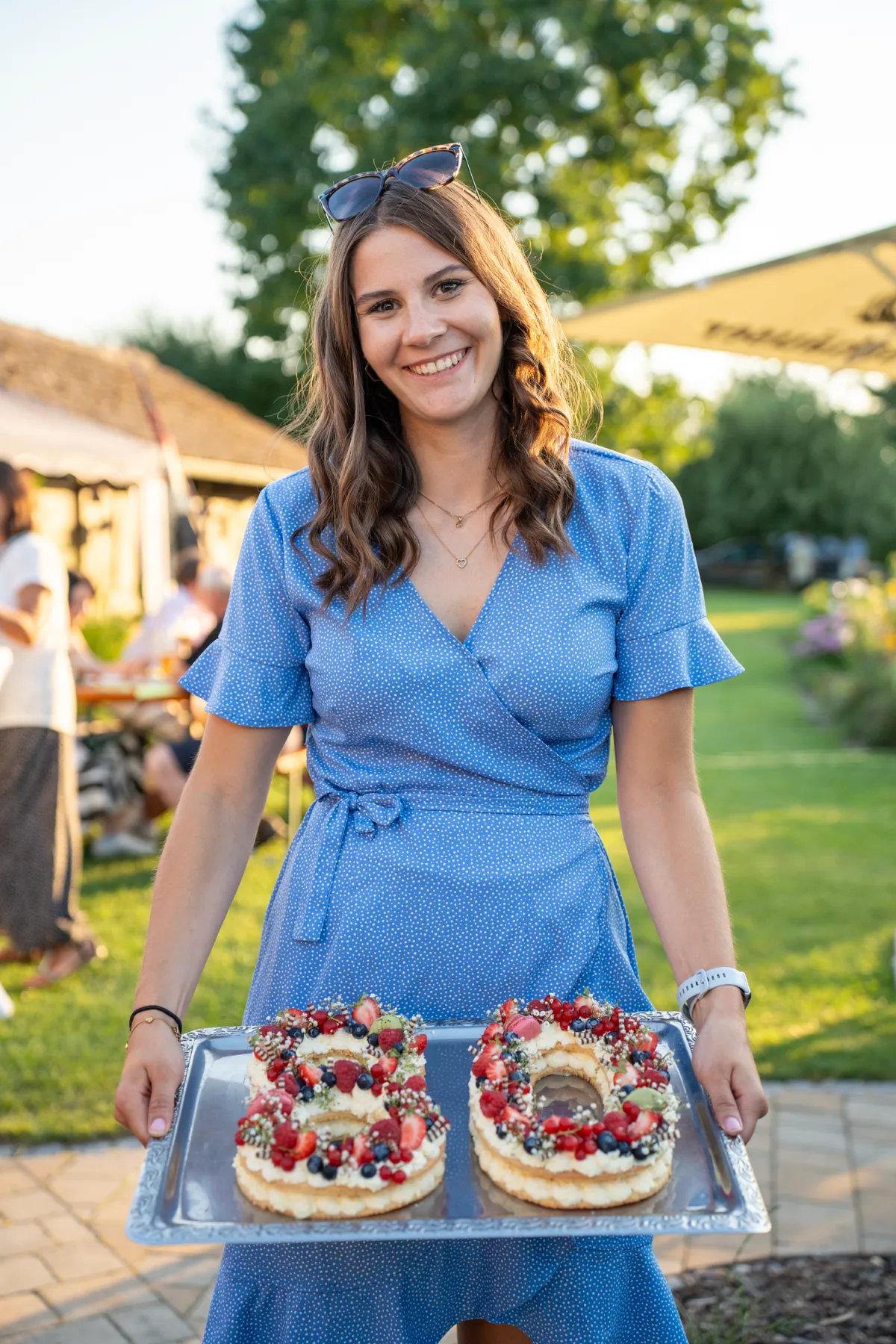 Eine Frau im blauen Kleid steht lächelnd mit dem Tortentablett im Garten.