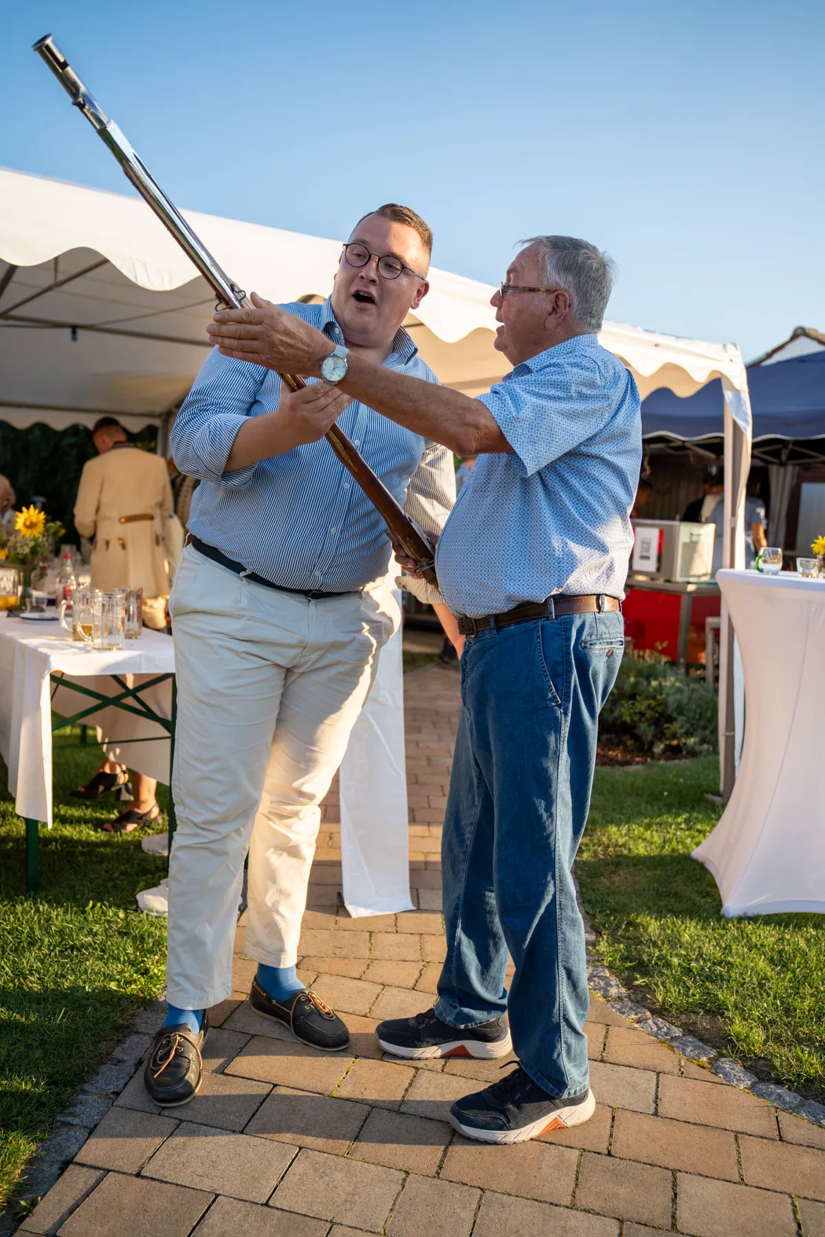 Der jüngere Mann hält das Gewehr, der ältere zeigt ihm etwas daran, beide lächeln.