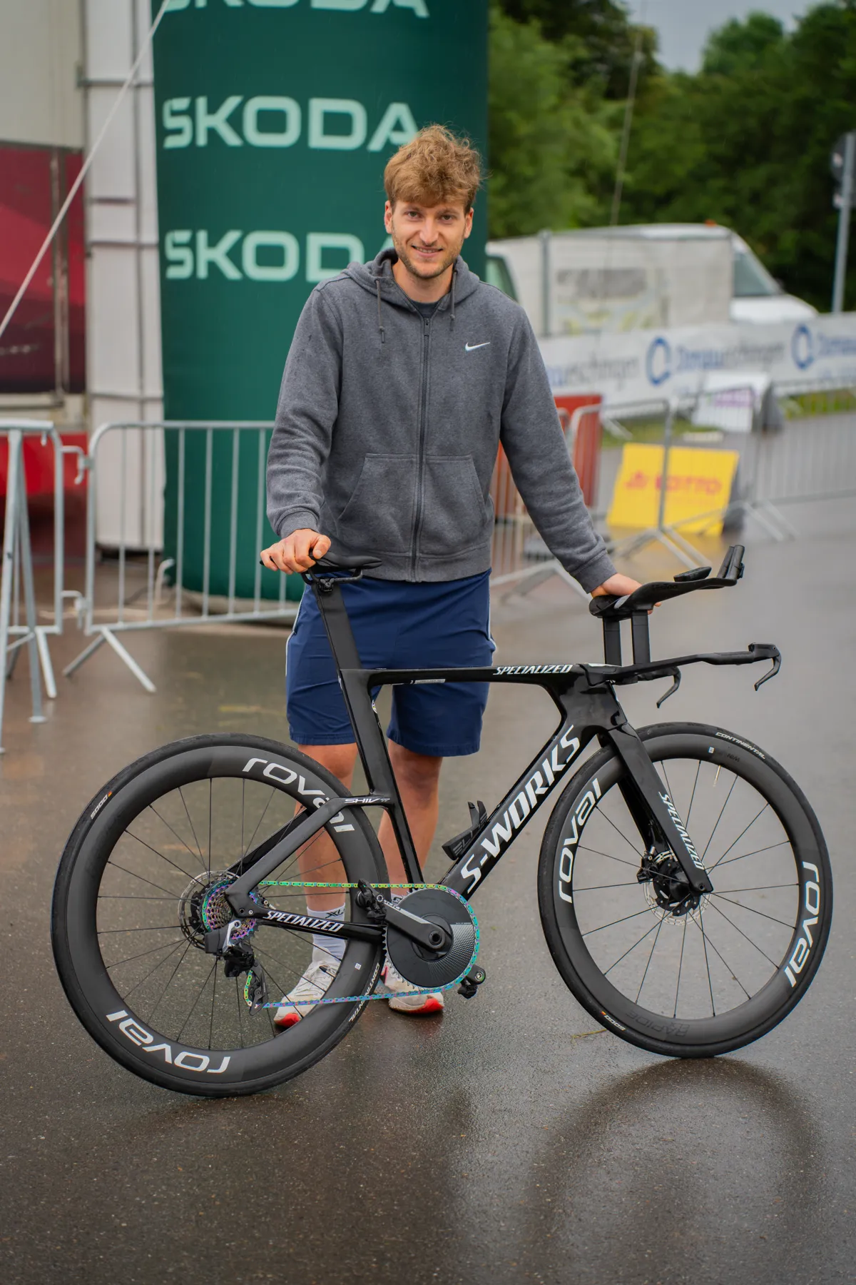 Mann steht mit Rennrad auf nassem Asphalt neben Werbebannern.