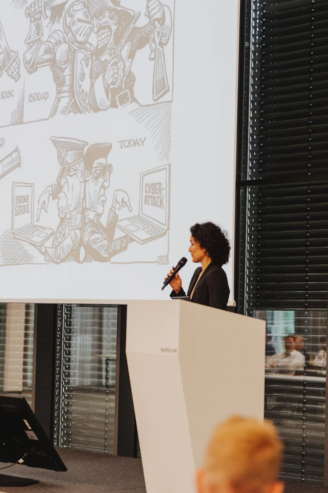 Rednerin mit Mikrofon am Pult vor einer Leinwand mit Karikatur zu Cyber-Angriffen.