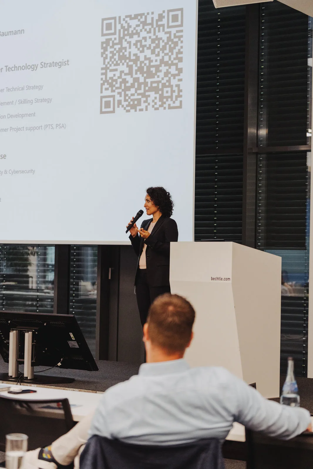 Eine Rednerin spricht mit Mikrofon neben dem Pult, auf der Leinwand eine Folie mit QR-Code.