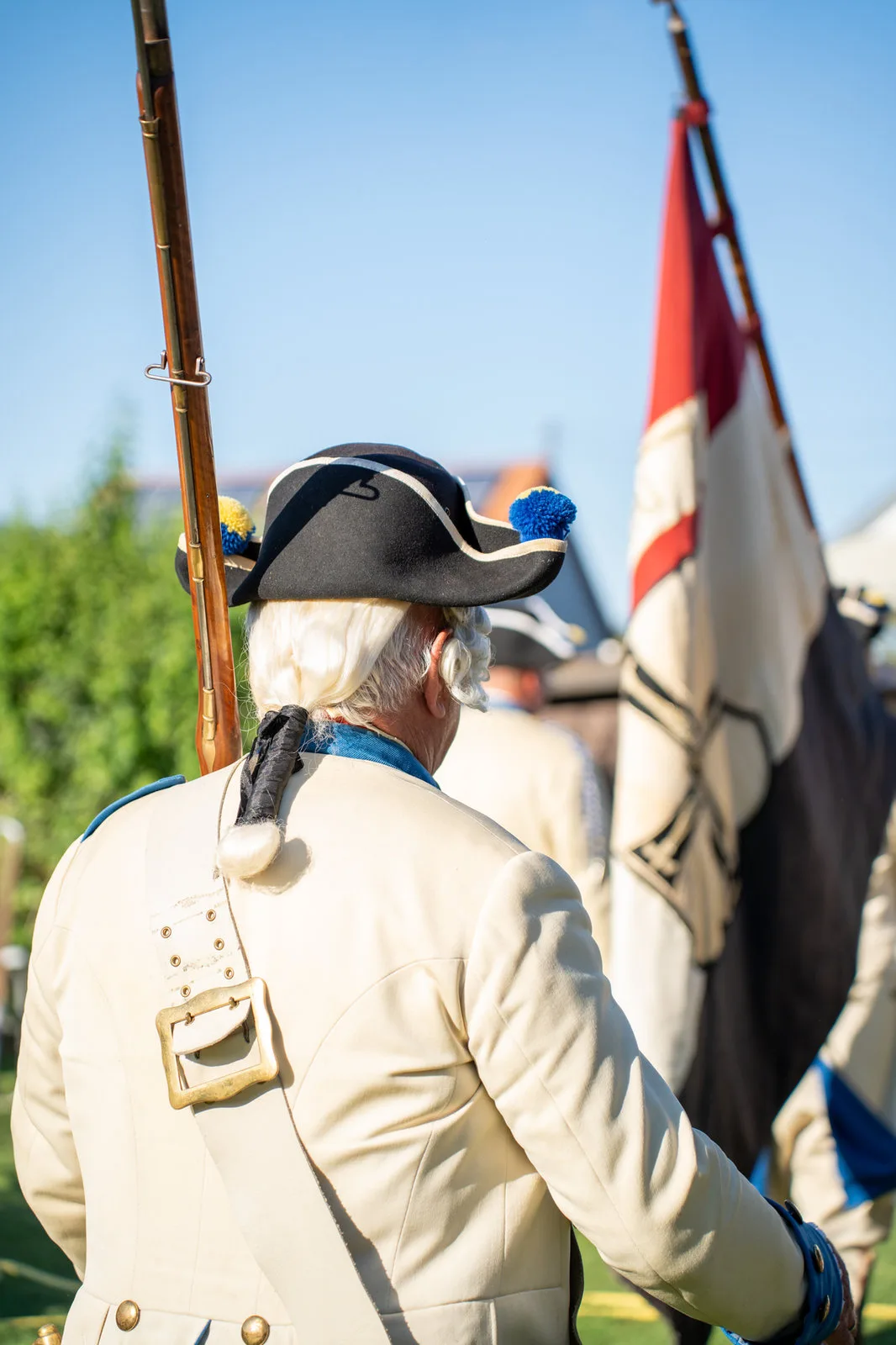 Gardist in historischer Uniform mit Dreispitz und geschulterter Muskete von hinten, daneben eine Fahne.