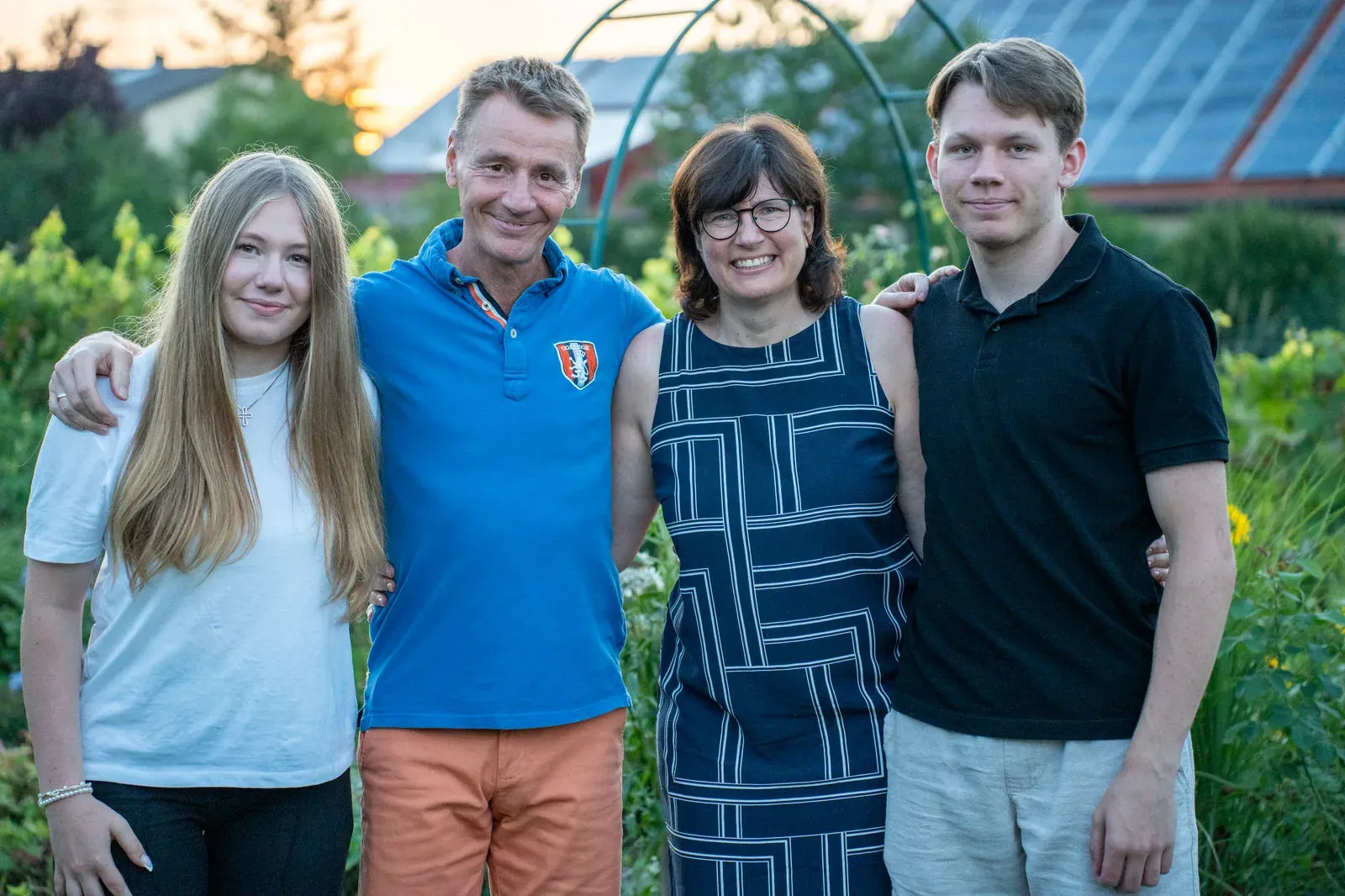 Vier Personen stehen Arm in Arm in einem Garten im Abendlicht, dahinter ein Gewächshaus.