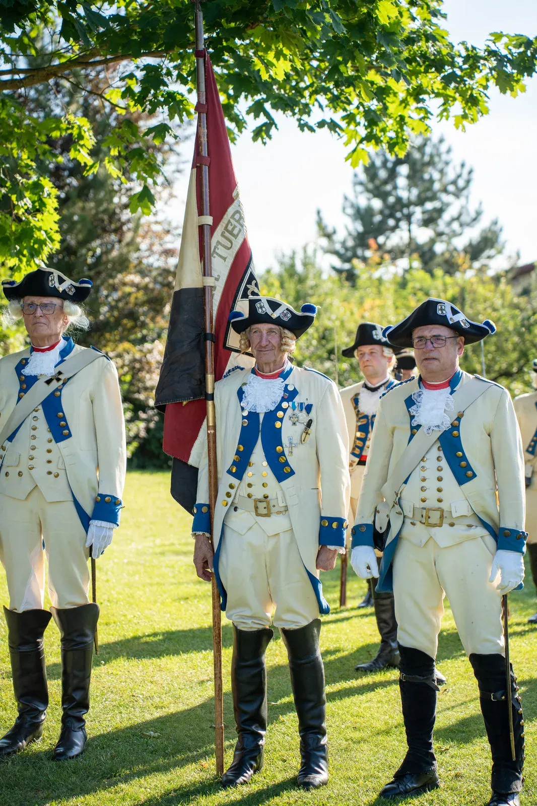 Drei Gardisten in weiß-blauen Uniformen, in der Mitte der Fahnenträger mit der Standarte.