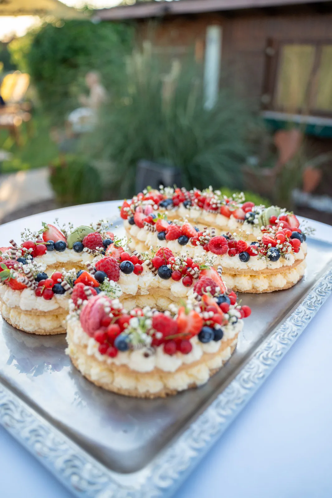 Drei mit Beeren, Macarons und Blüten verzierte Torten stehen auf einem Silbertablett im Garten.