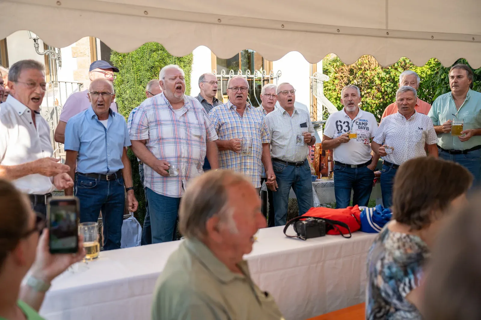 Ein Männerchor singt beim Gartenfest unter dem Zeltdach, einige halten Biergläser.
