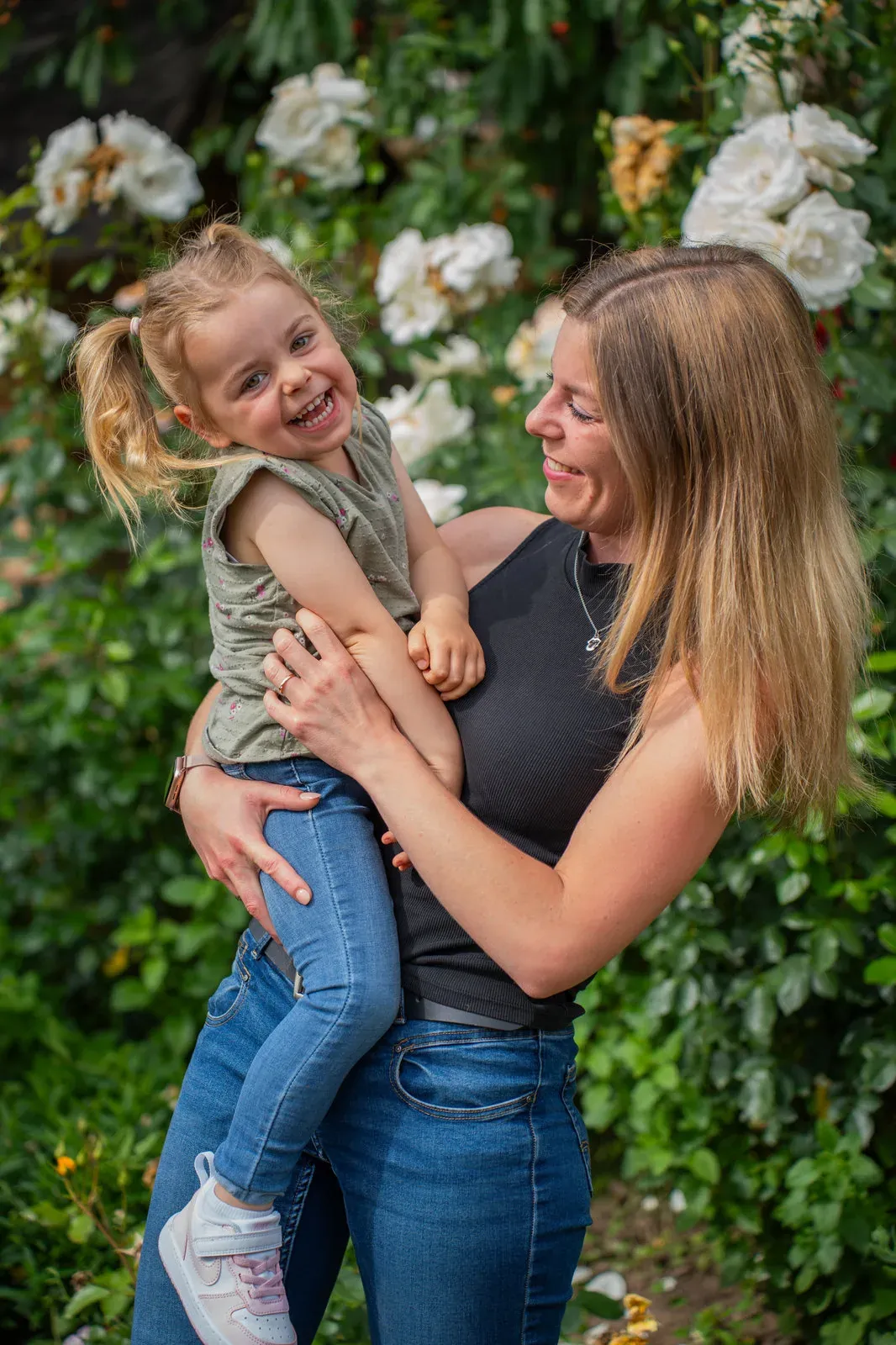 Eine Mutter hält ihre lachende Tochter auf dem Arm, im Hintergrund blühen weiße Rosen.