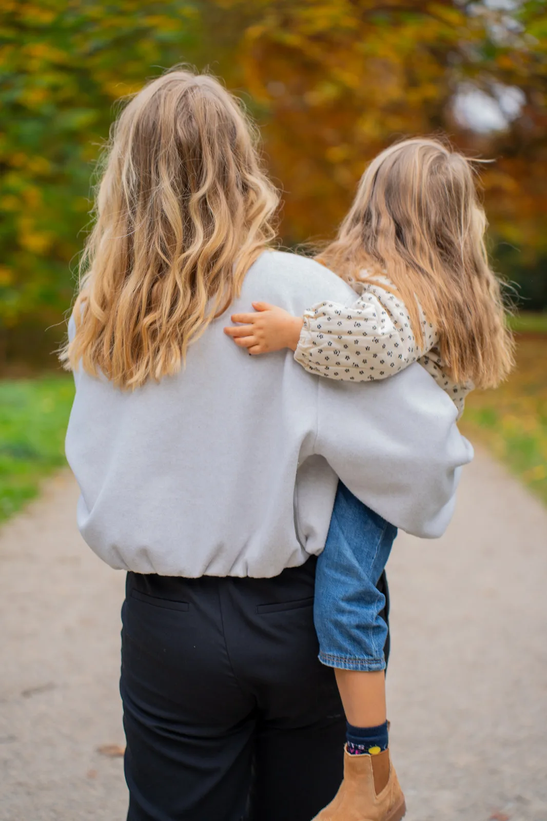 Rückansicht: Eine Frau trägt ein Kind auf dem Arm einen herbstlichen Weg entlang.