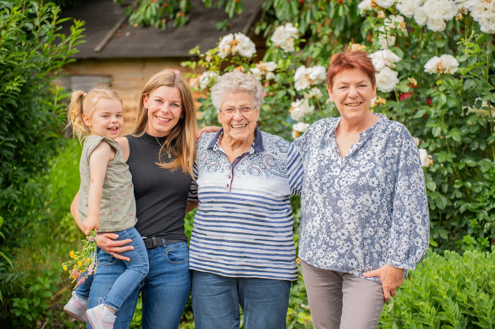 Familienportrait im Rosengarten: Urgroßmutter, Großmutter, Mutter und Kind lachen in die Kamera.