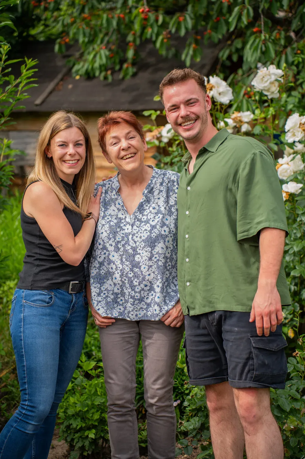 Mutter mit erwachsener Tochter und erwachsenem Sohn vor blühenden Rosenbüschen.