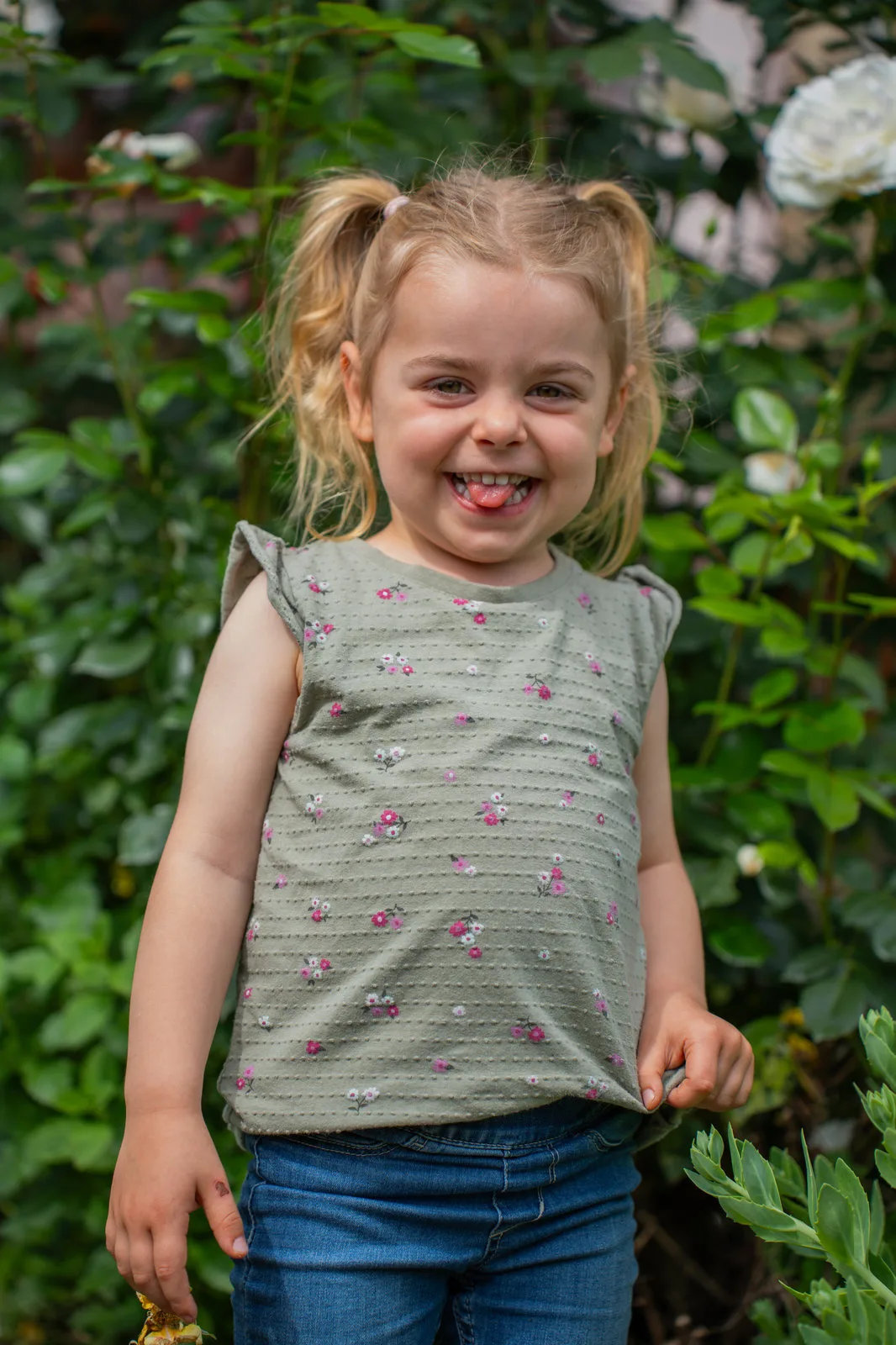 Übermütiges Kinderportrait zwischen Rosenbüschen: Das Mädchen streckt vergnügt die Zunge heraus.