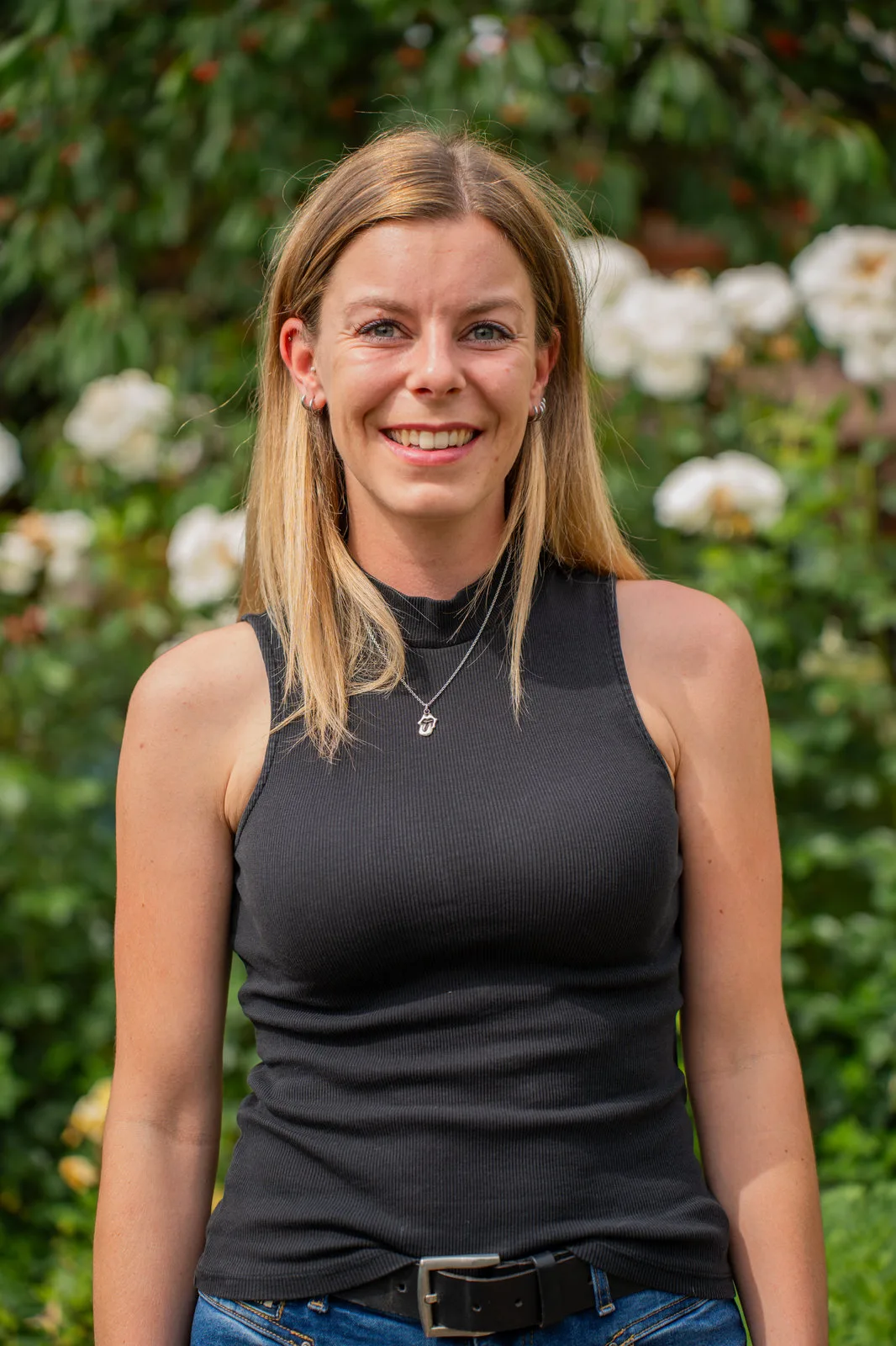 Lächelnde junge Frau mit langen blonden Haaren vor einer Rosenhecke.