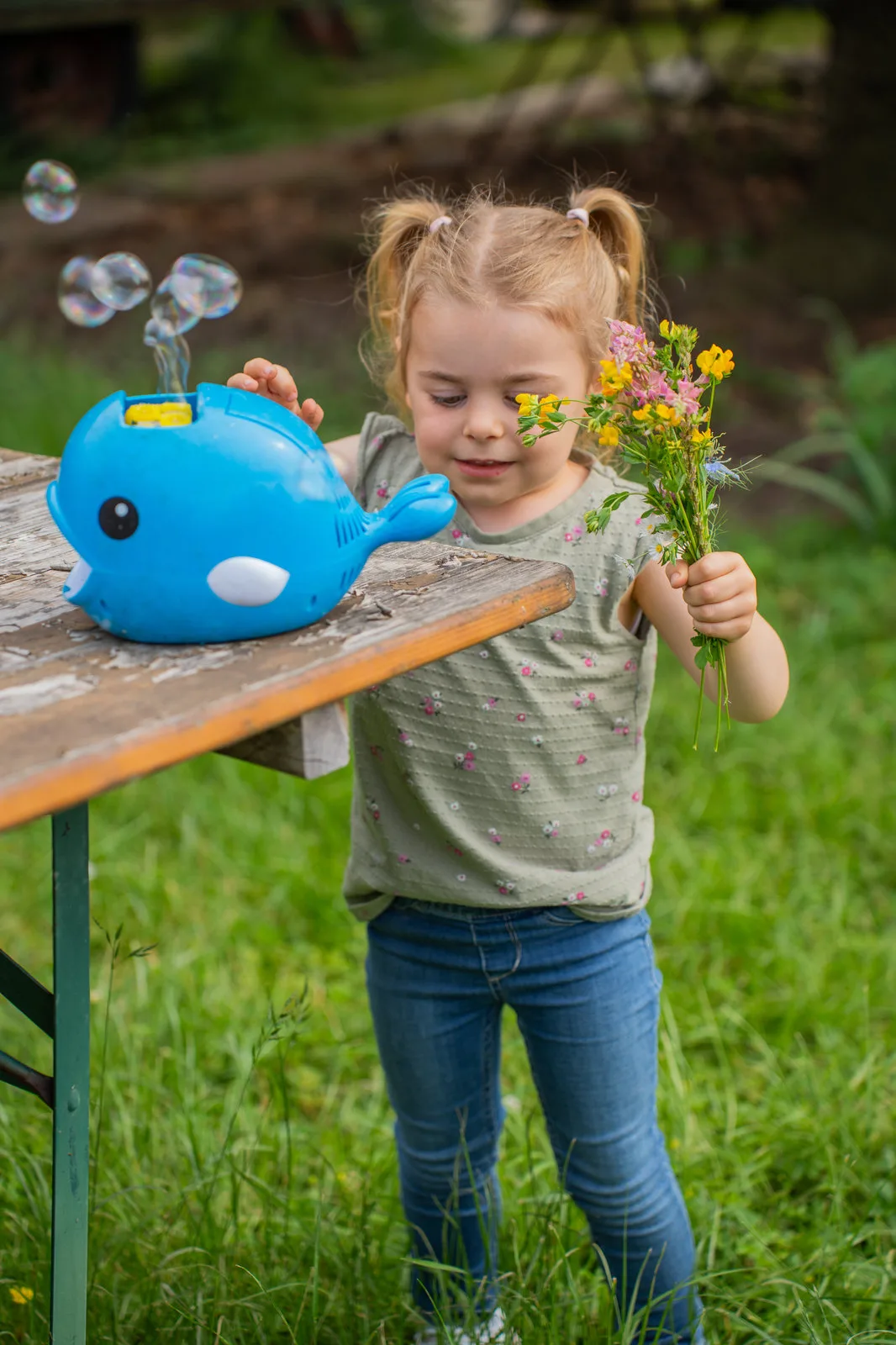 Ein Kind betrachtet eine walförmige Seifenblasenmaschine auf dem Gartentisch, Blumenstrauß in der Hand.
