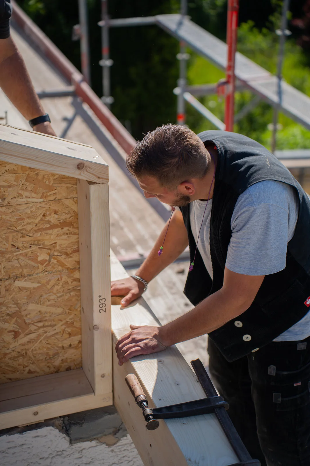 Ein Zimmermann kontrolliert ein Holzelement auf der Baustelle, im Hintergrund das Gerüst.