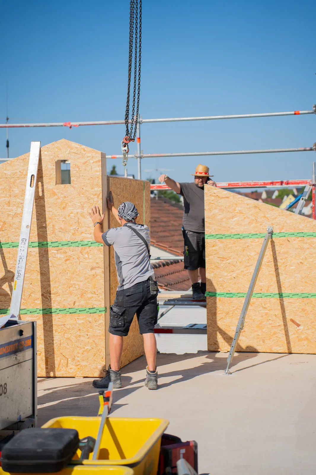 Zimmerleute stellen am Kran hängende Holzwandelemente auf dem Rohbau auf.