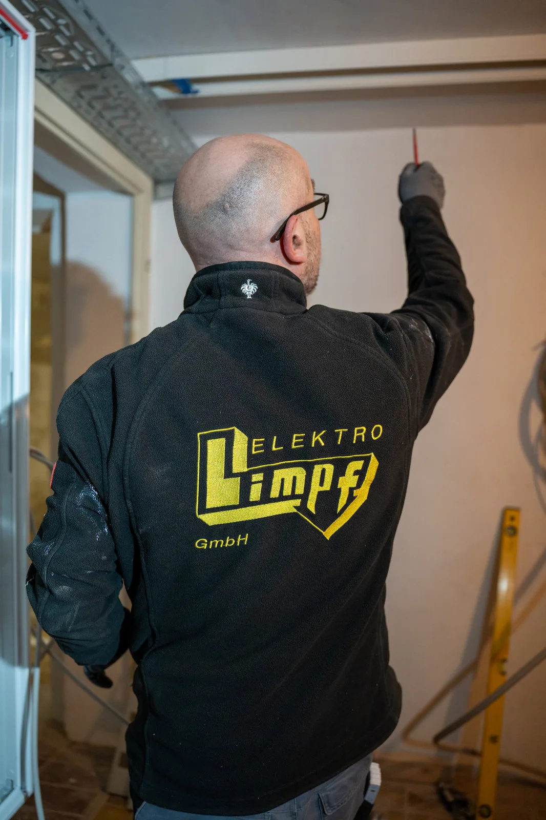 Ein Elektriker mit Firmenjacke arbeitet an der Wandinstallation eines Rohbaus.