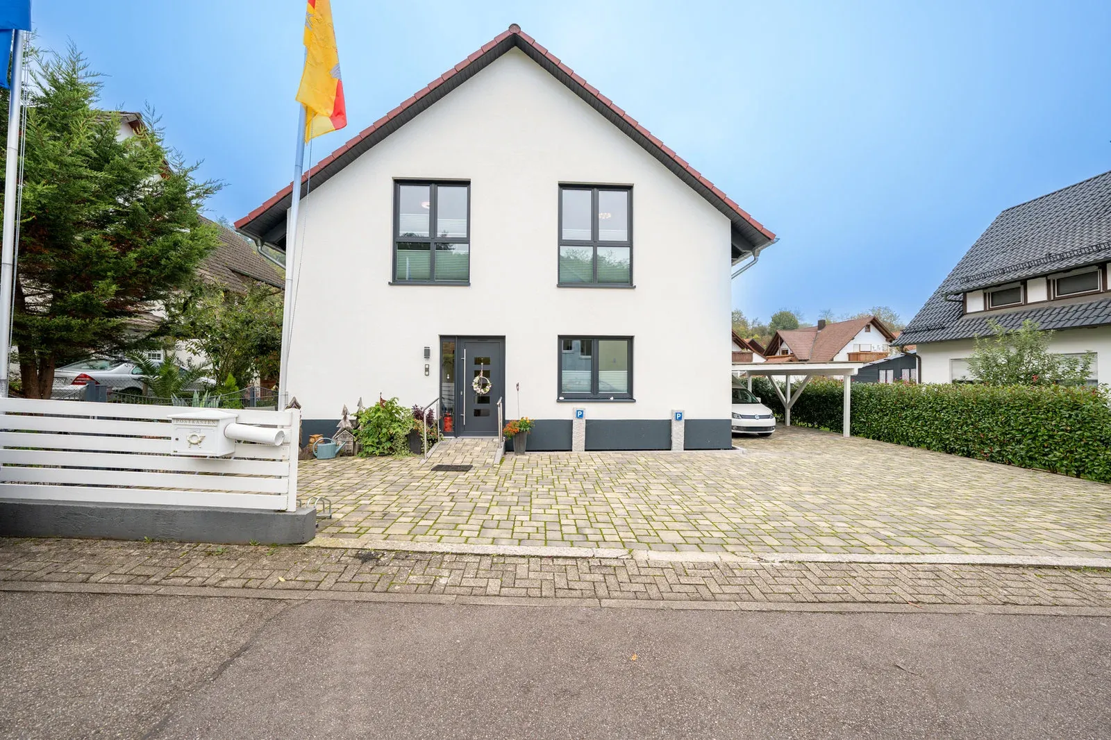 Frontansicht des weißen Giebelhauses mit gepflasterten Stellplätzen und Fahnenmast.