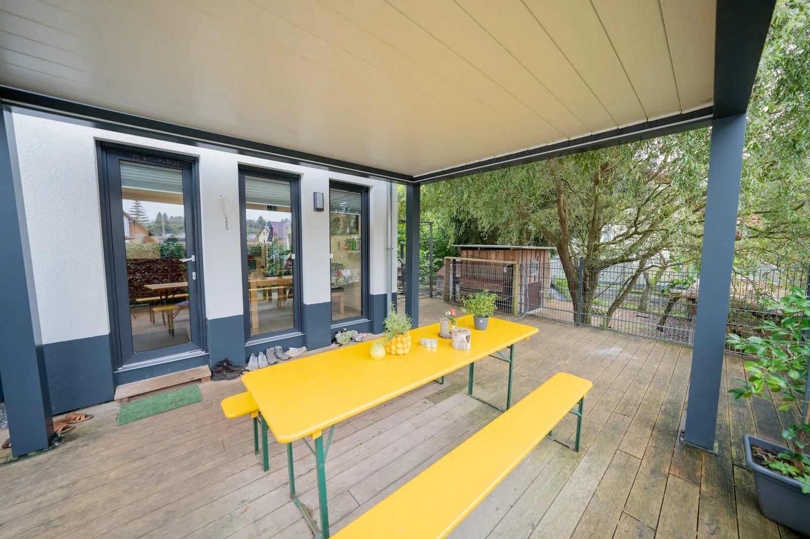 Überdachte Holzterrasse mit leuchtend gelber Bierzeltgarnitur und Gartenblick.