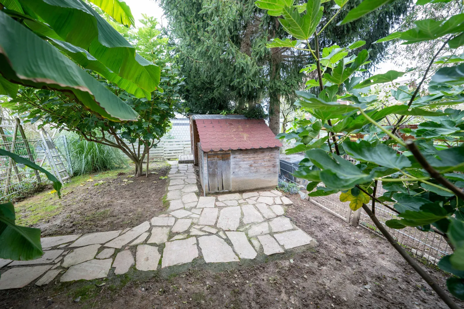 Ein Natursteinweg führt durch den Garten zu einem kleinen Holzschuppen mit rotem Schindeldach.