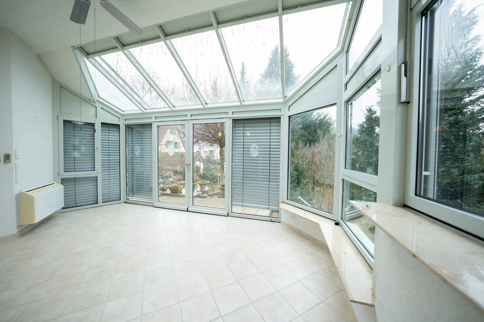 Heller Wintergarten mit Glasdach, Jalousien und Blick auf die Terrasse.