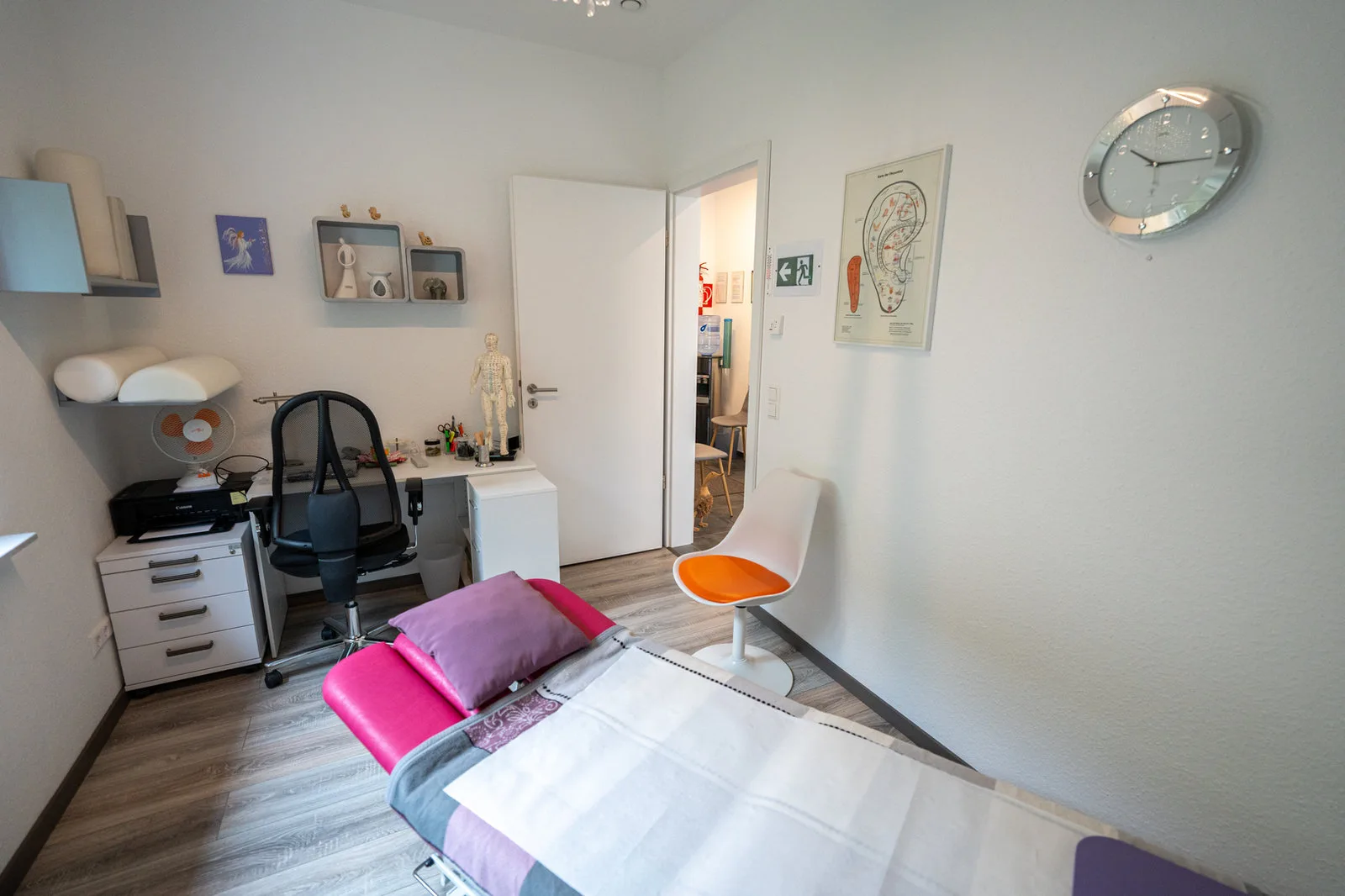 Behandlungszimmer einer Praxis mit Liege, Schreibtisch und Ohrakupunktur-Tafel.