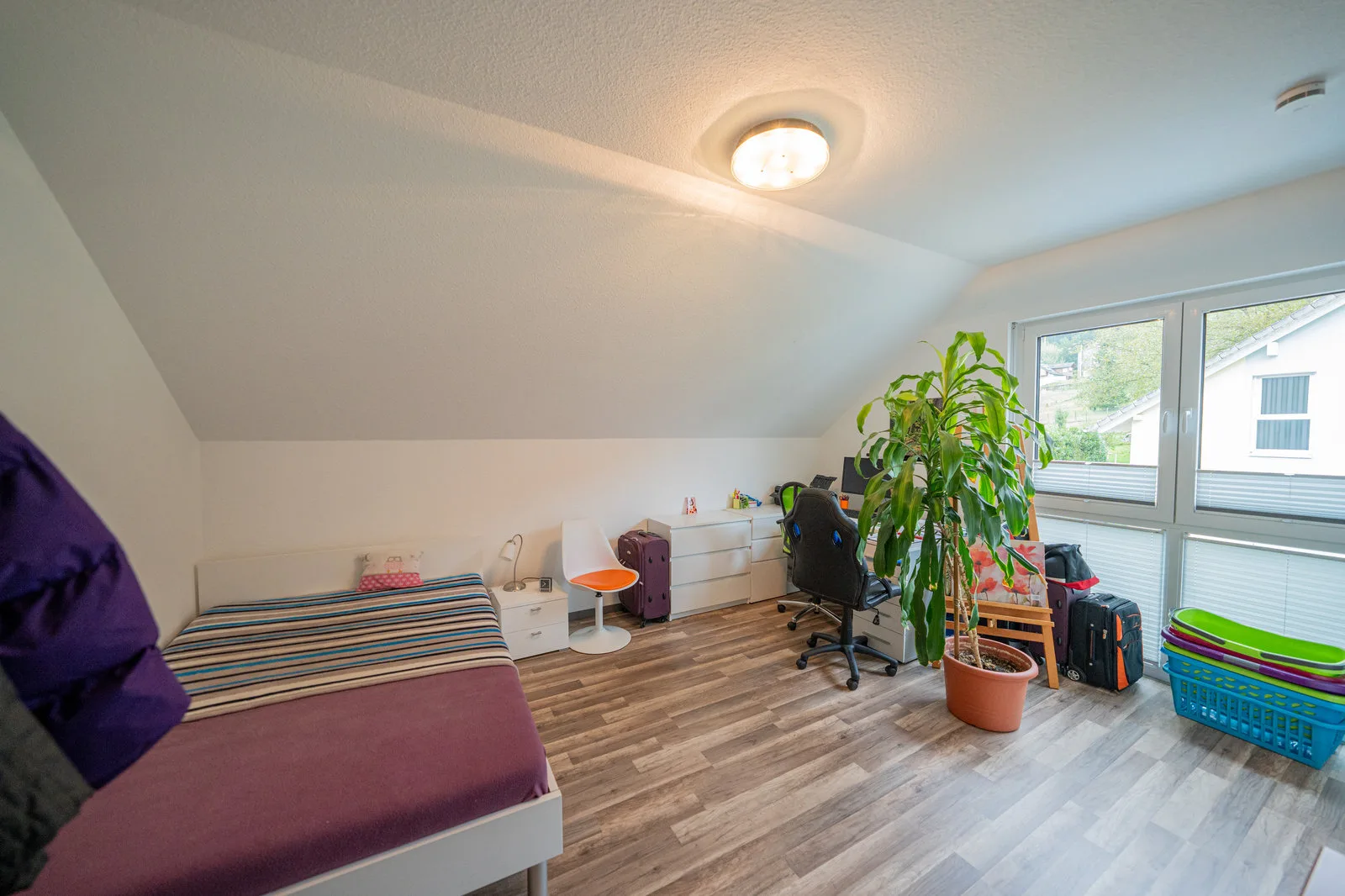 Dachgeschosszimmer mit Bett, Schreibtisch und großer Zimmerpflanze am Fenster.