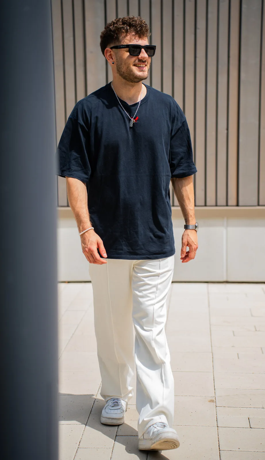 Streetstyle-Portrait: Ein junger Mann mit Sonnenbrille und Kette läuft lässig an einer Lamellenfassade vorbei.