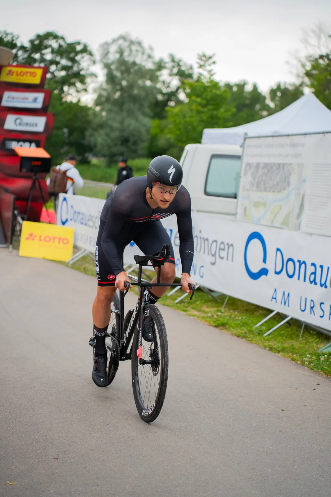 Ein Zeitfahrer tritt auf seinem Rennrad kraftvoll in die Pedale, im Hintergrund Sponsorenbanner.