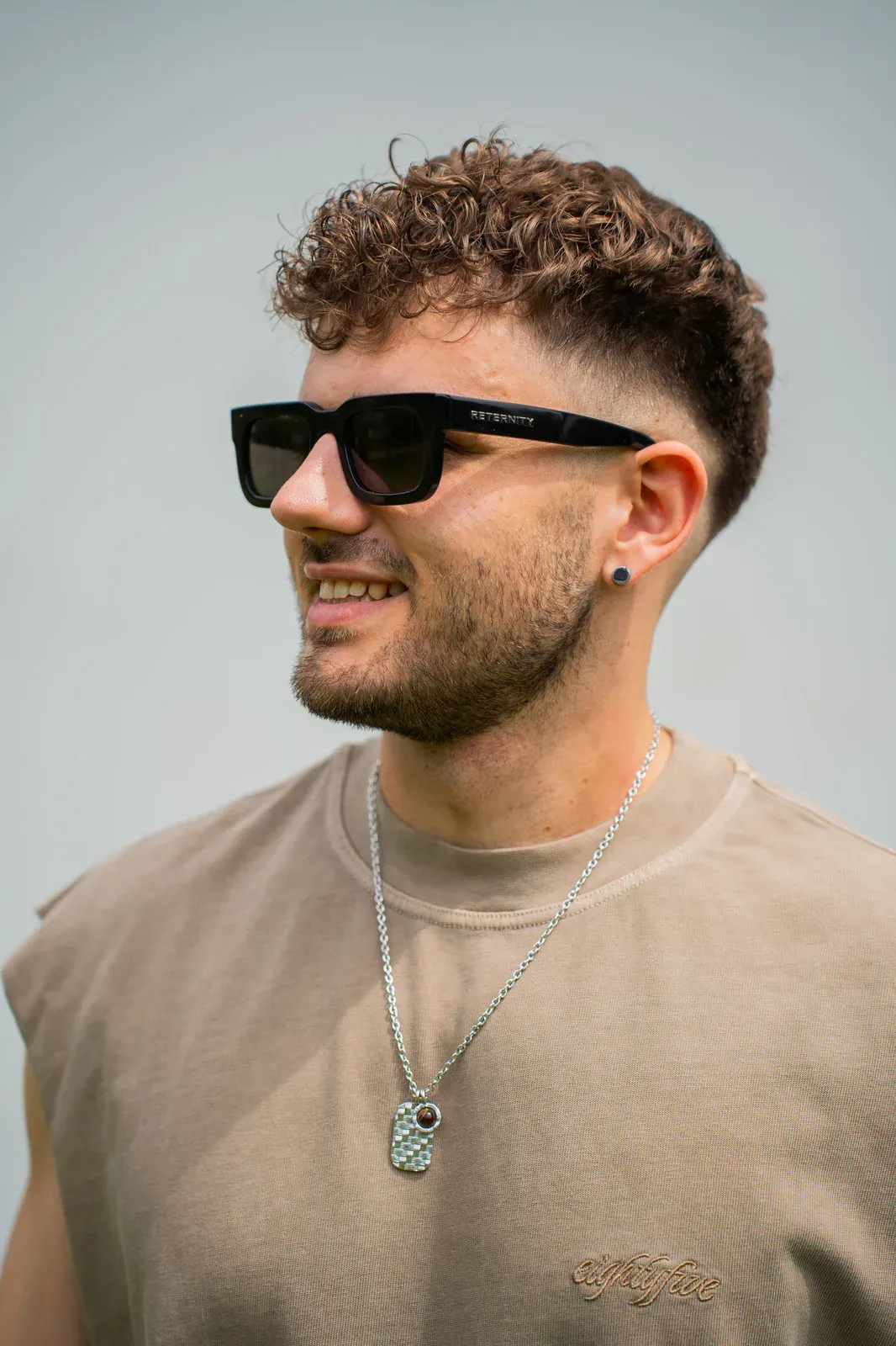 Seitliches Portrait eines jungen Mannes mit Locken, Sonnenbrille und Silberkette im beigen Shirt.