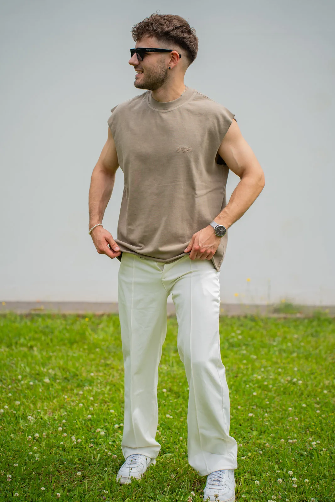 Ein junger Mann mit Sonnenbrille posiert lässig im beigen Muskelshirt und weißer Hose.